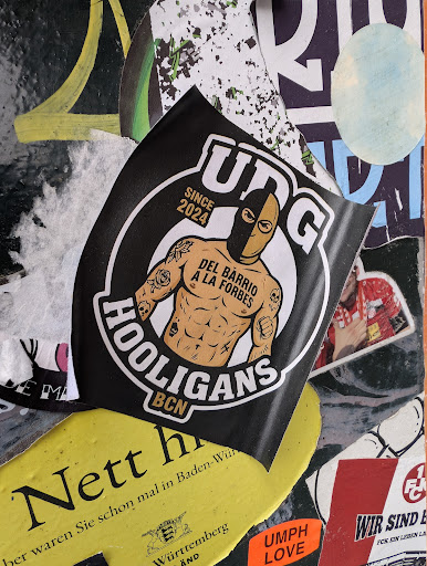 This sticker features a black and white design with a character wearing a half-black mask. The text "UNG SINCE 2024" appears at the top. Below the character is the phrase "DEL BARRIO A LA FORBES" and "HOOLIGANS BCN" at the bottom, suggesting a connection to the city of Barcelona.