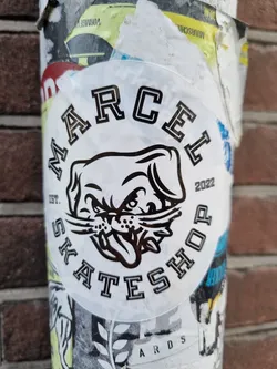 Street sticker A round sticker with a black and white illustration of a dog's head and the text "MARCEL SKATESHOP" around it. The year 2022 is printed on the right side of the circle. 
