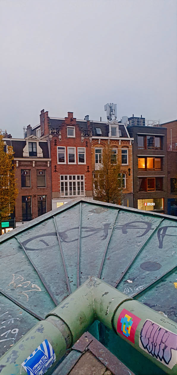 The image shows a metal pipe with several stickers affixed to it. The background features buildings, creating an urban atmosphere. One sticker is particularly visible, displaying a pixelated cross design on a pink background.