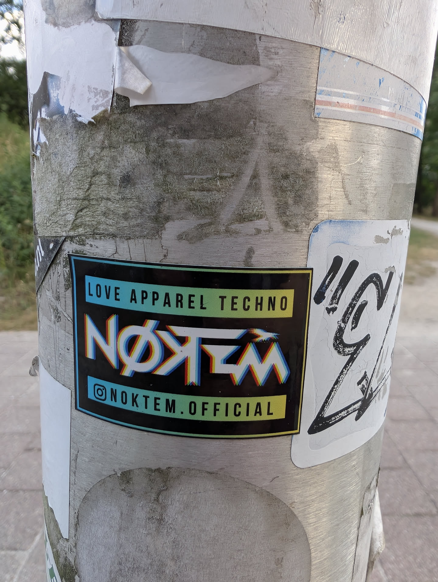 A black rectangular sticker with a gradient border. It features 'LOVE APPAREL TECHNO' at the top and '@NOKTEM.OFFICIAL' at the bottom on blue-to-yellow gradient bars. The center displays 'NØKTEM' in a stylized white font with a red and blue chromatic aberration effect.