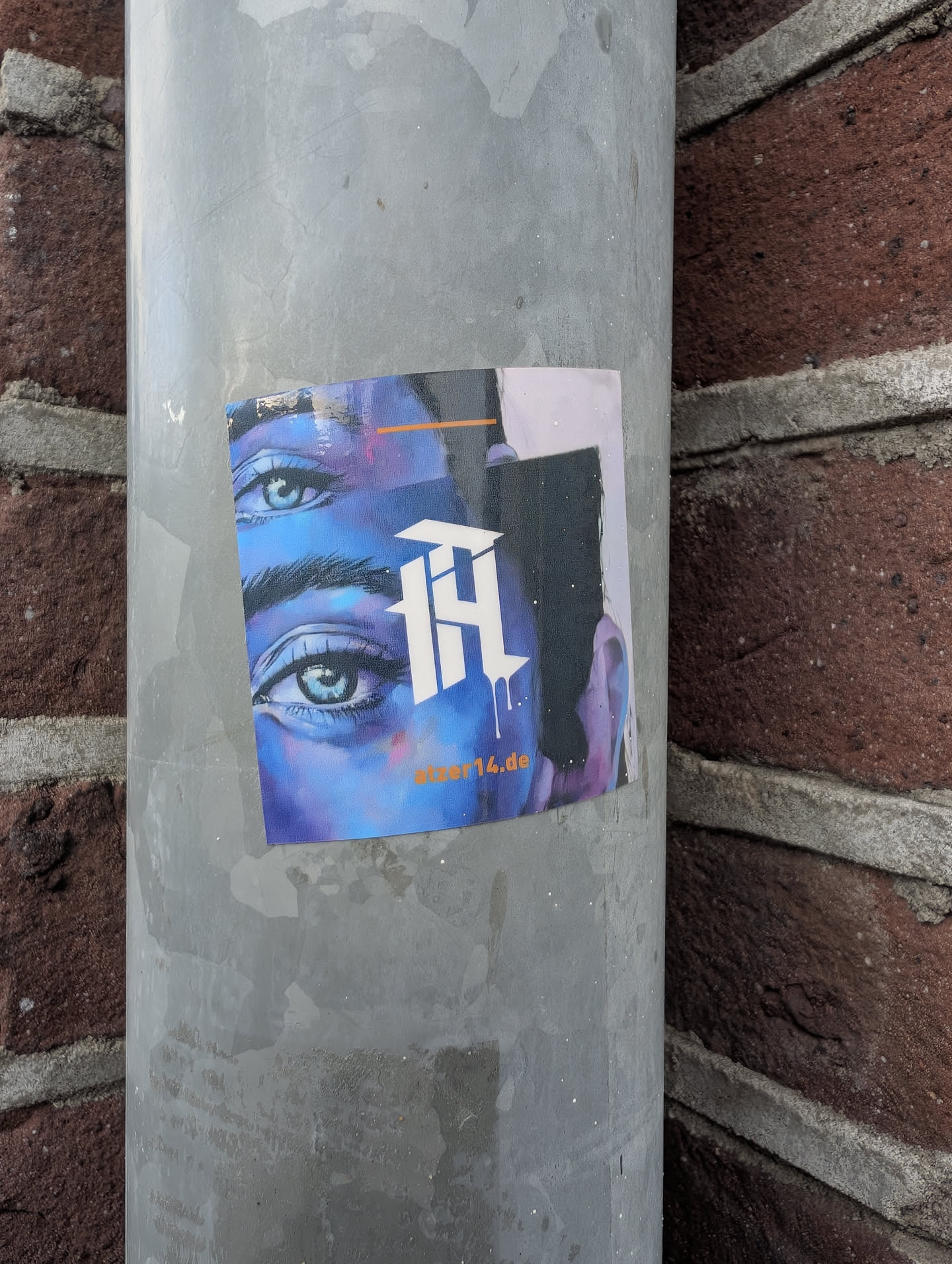 Square sticker depicting a face in blue and purple tones with detailed eyes. Features a central white gothic monogram and the orange text 'atzer14.de' at the bottom.