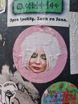 Street sticker This is a close-up image of a round sticker featuring a woman with white hair and striking green eyeshadow.  Her expression is serene, with her eyes closed. The sticker is predominantly pink. A website address, www.neverever.nl, is visible at the bottom.  Surrounding stickers are partially visible, one of which is a Spotify code and another referencing "Virtual Virality, Online Intimacy & Digital Detox."