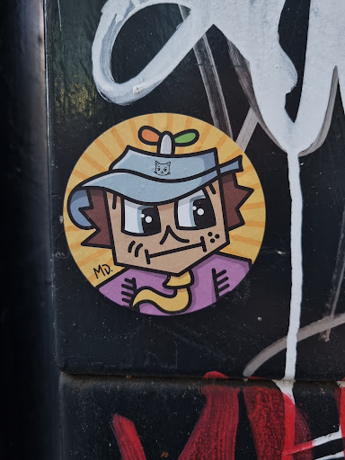 A round sticker featuring a cartoon character with a hat and a small plant on its head. The character has a simple, almost pixelated style and is wearing a purple shirt. The background of the sticker is yellow with radiating lines. The sticker appears to be affixed to a dark surface with other graffiti present.