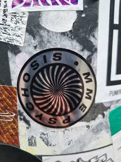 Street sticker A round, silver sticker with a black and white hypnotic spiral design. The words \"PSYCHOSIS MEME\" are written around the spiral in a circular pattern. The sticker appears to be affixed to a surface with other stickers and graffiti.