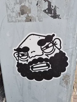 Black and white sticker of a man with a beard. He has a grumpy expression and seems to be scowling. The style is simple and cartoonish.