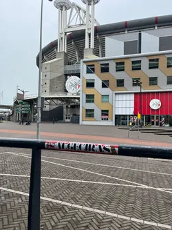 Street sticker The sticker is rectangular, predominantly red and white, with black accents. On the left side, there is a detailed crest-like emblem in black and white, resembling a shield with a castle or building above waves, enclosed within a circular border. To the right of the crest, the word 'ALMECIRAS' is written in bold, white capital letters against a solid red background. The sticker shows some minor wear and tear, particularly at its edges where it adheres to the black metal railing.