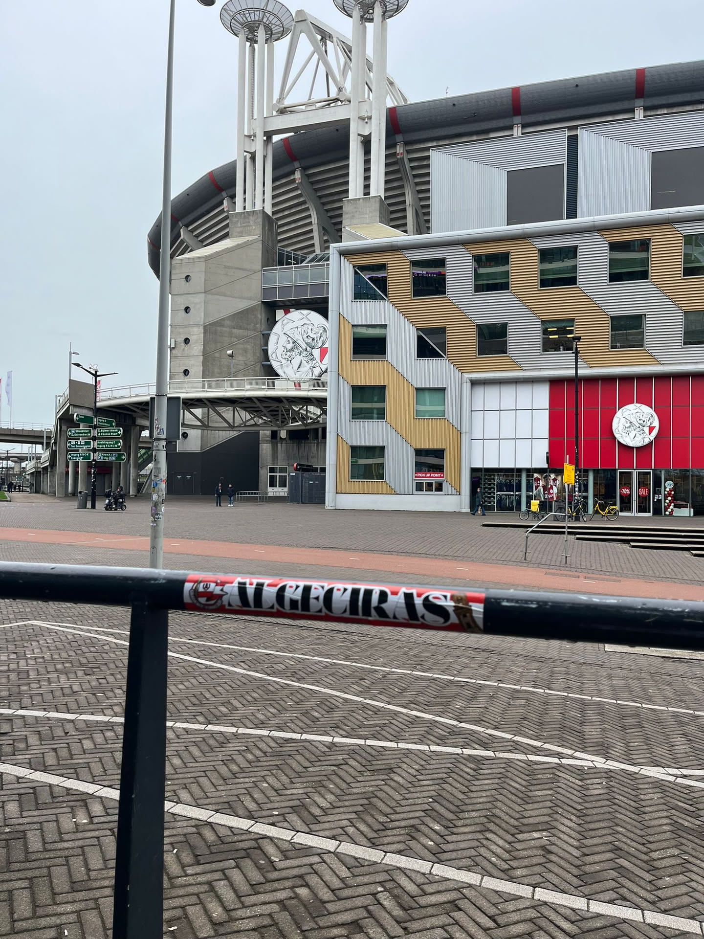 The sticker is rectangular, predominantly red and white, with black accents. On the left side, there is a detailed crest-like emblem in black and white, resembling a shield with a castle or building above waves, enclosed within a circular border. To the right of the crest, the word 'ALMECIRAS' is written in bold, white capital letters against a solid red background. The sticker shows some minor wear and tear, particularly at its edges where it adheres to the black metal railing.