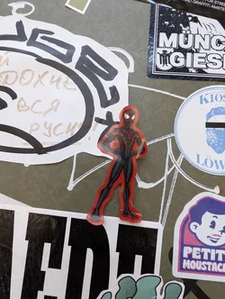 A vinyl sticker featuring a stylized image of Spider-Man in a red and black suit. The sticker is placed on a surface with various other stickers and graffiti.