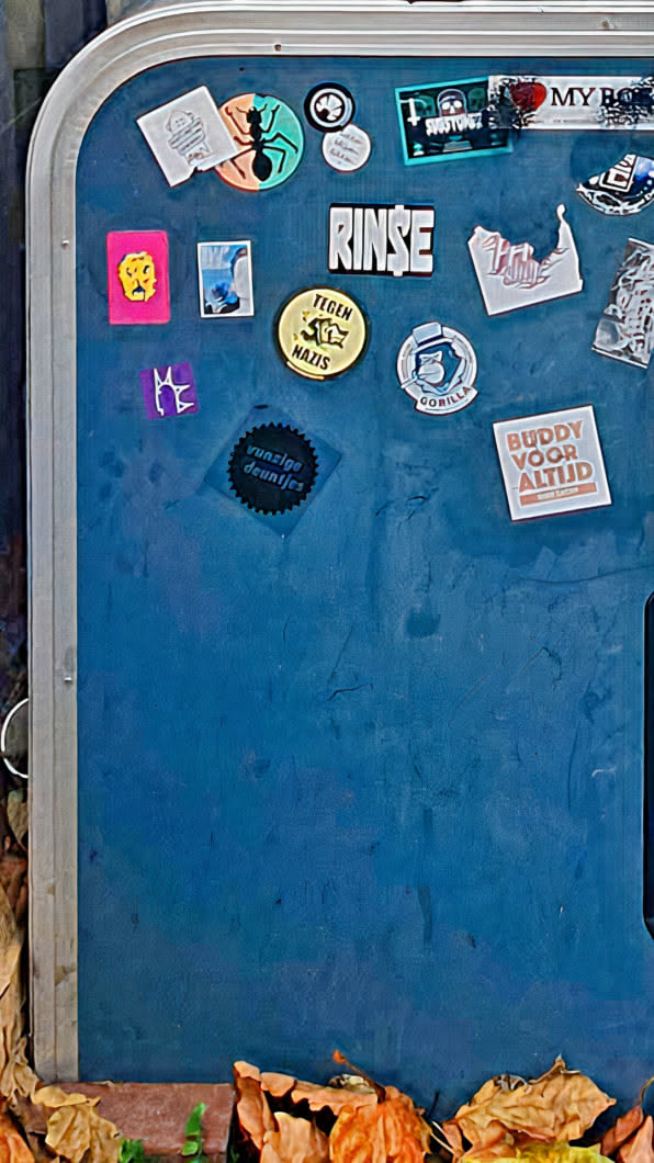 The image features a blue surface adorned with various stickers, creating a collage-like effect. The stickers showcase a range of designs, colors, and themes. A particular sticker shows a black cogwheel with the text 'vunzige deuntjes' inside. The presence of leaves at the bottom suggests an outdoor setting, possibly a public fixture or wall.