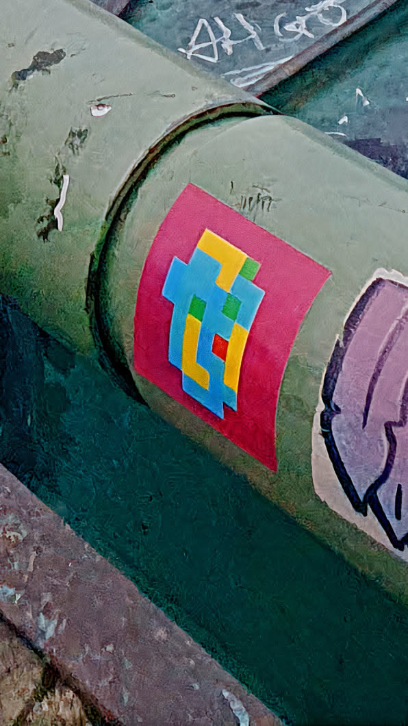 A sticker with a pink background with a pixelated creature in blue, yellow, green and red is stuck on a green painted pipe.