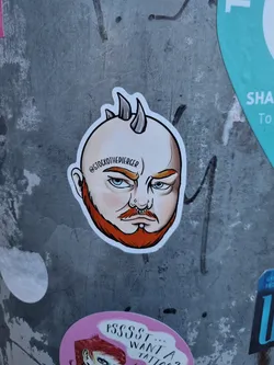 Close-up of a sticker featuring a stylized illustration of a man with short horns, a beard, and facial piercings. The art style is reminiscent of cartoonish tattoo flash.  The Instagram handle '@stocktothepiercer' is visible on the sticker.