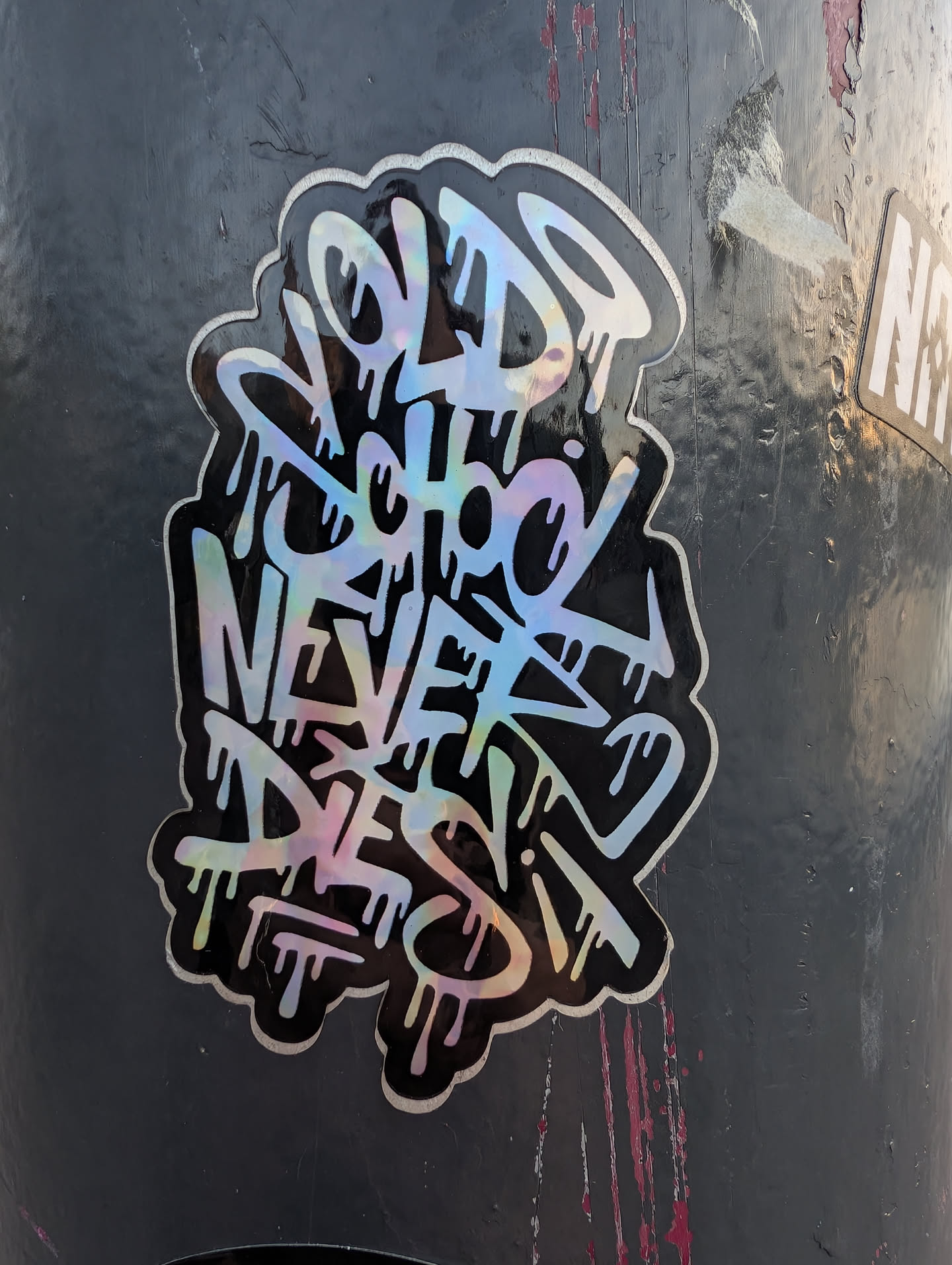 A die-cut sticker featuring graffiti handstyle lettering with a dripping effect. The text 'OLD SCHOOL NEVER DIES' is rendered in an iridescent, color-shifting material against a black background with a thin silver border.