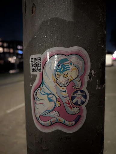 A sticker featuring a white tiger with blue stripes and a pink outline, adhered to a pole. The tiger has a distinctive, stylized look, and a QR code is visible on the side. Text on the sticker includes 'kl club.cool' and 'karate dojo'.