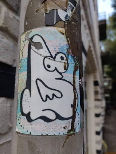 A charming sticker featuring a simple, cartoon-like creature in black and white with a pastel background. The sticker shows some wear, suggesting it has been exposed to the elements. It is affixed to a weathered pillar or pole, adding to its urban aesthetic.