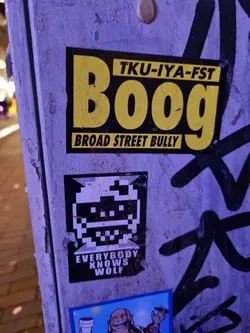 Street sticker A yellow and black sticker with the text "Tku-iya-fst Boog" and below it "Broad Street Bully". There is also a smaller pixel art sticker underneath it with the text "Everybody Knows Wolf".