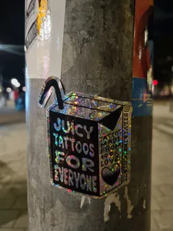 Street sticker Holographic sticker featuring a juice box design with the text "Juicy Tattoos for Everyone."  The sticker also has smaller text that reads "Tattoos made from 100% love." A heart is included in the design. The artist's Instagram handle is visible on the sticker.