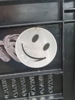 A simple, round sticker with a slightly mischievous smiley face design.  The sticker is white or light gray with black features and appears to be applied to a dark gray or black plastic surface.