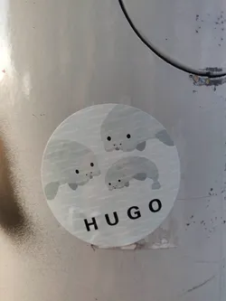 Street sticker A round sticker featuring an illustration of three adorable cartoon manatees swimming. The word "HUGO" is printed underneath the manatees. The sticker appears to be affixed to a light gray surface.