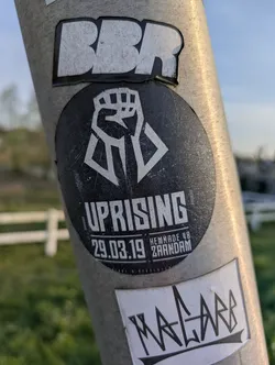 This is a round sticker with a black background and white text and graphics. The main image is a raised fist with a geometric design below it. The text "UPRISING" is prominently displayed, followed by "29.03.19 HEMKADE 48 ZAANDAM", indicating a date and location, likely for an event.