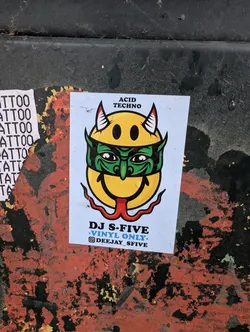 Street sticker The sticker features a yellow smiley face with devilish horns and a green mask over its eyes. A red tongue is sticking out and curved to the sides. Above the smiley, it reads 'ACID TECHNO' in black capital letters. Below the smiley, it says 'DJ S-FIVE' in blue, followed by 'VINYL ONLY.' in smaller blue text. Beneath that, an Instagram logo is followed by the handle '@DEEJAY_SFIVE'. The sticker has a white background and appears to be slightly creased.