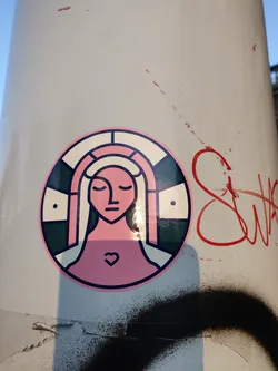 Close-up view of a round sticker featuring a minimalist illustration of a woman's face with a heart on her chest. The style is geometric with a color palette of pink, cream, and dark green. The sticker is affixed to a light beige surface with some graffiti visible nearby.
