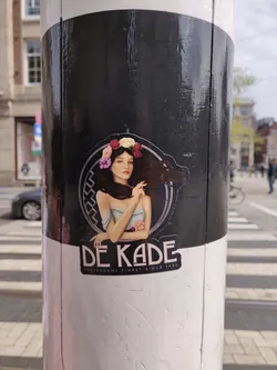 Sticker featuring a stylized illustration of a woman with flowers in her hair. The sticker is black and white with pink accents. It also contains the text 'DE KADE' and 'AMSTERDAMS FINEST SINCE 1986'.