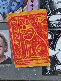 A rectangular sticker featuring a stylized illustration of a cat in red and orange, reminiscent of ancient Egyptian art. The cat is depicted sitting, and the design incorporates hieroglyphic-like symbols.