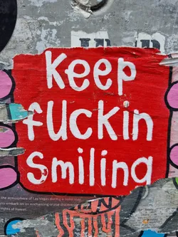 A red sticker with white hand-painted text. The sticker is slightly damaged and torn at the edges. It's affixed to a wall that shows other layers of stickers and posters underneath.