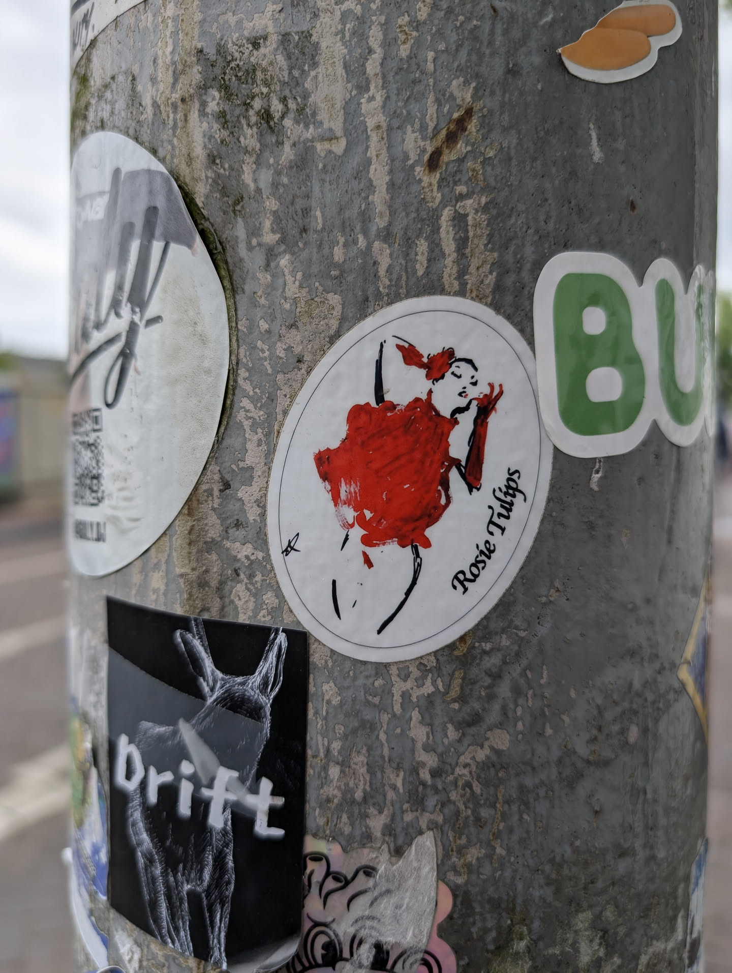 Street sticker The sticker is circular and white with a black outline. It depicts a minimalist illustration of a female dancer in a dynamic pose. The dancer is rendered in black lines, with her hair tied up and wearing long black gloves. She is adorned in a voluminous red dress, suggested by broad brushstrokes. To the right of the figure, in elegant script, is the text 'Rosie Tulips'. The overall style is artistic and expressive.