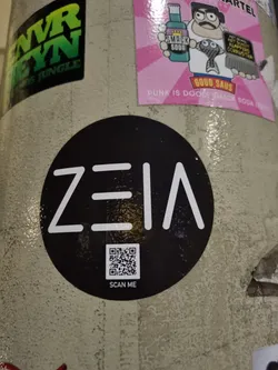 Black and white circular sticker with the word "ZEIA" written in a stylized font.  It includes a QR code with the text "SCAN ME" underneath.