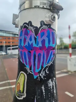 A vibrant sticker featuring the phrase "Hard to Love" in stylized lettering, encased within a heart shape and framed by barbed wire. The color scheme is predominantly blue and pink, creating a striking visual contrast.