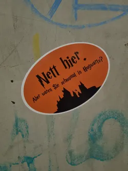 Street sticker An orange oval sticker featuring a silhouette of Hogwarts castle with the text "Nett hier. Aber waren Sie schonmal in Hogwarts?" which translates to "Nice here. But have you ever been to Hogwarts?" in German. The sticker is affixed to a grey concrete surface.