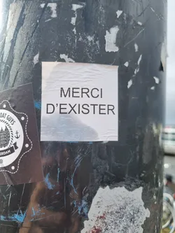 Street sticker A simple, yet impactful message of gratitude.  The sticker features the French phrase "Merci d'exister," which translates to "Thank you for existing." It is printed in a straightforward, sans-serif font on a white background.