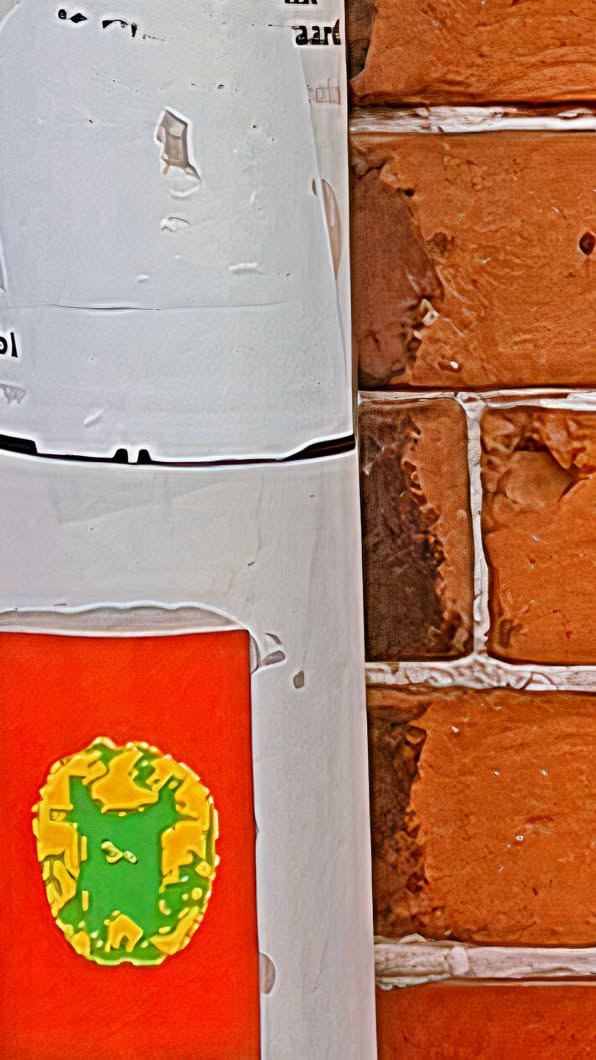 This image features a bright orange sticker with a green and yellow design, likely an artistic representation of a map or a symbol. The sticker is attached to a post, with a brick wall visible in the background, adding texture and depth to the image. The lighting is bright, highlighting the sticker's colors and the wall's details.