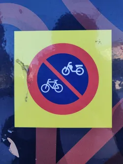 A yellow sticker with a red circle and a diagonal line through the images of a bicycle and a scooter.  The sticker is damaged, showing some peeling at the edges.