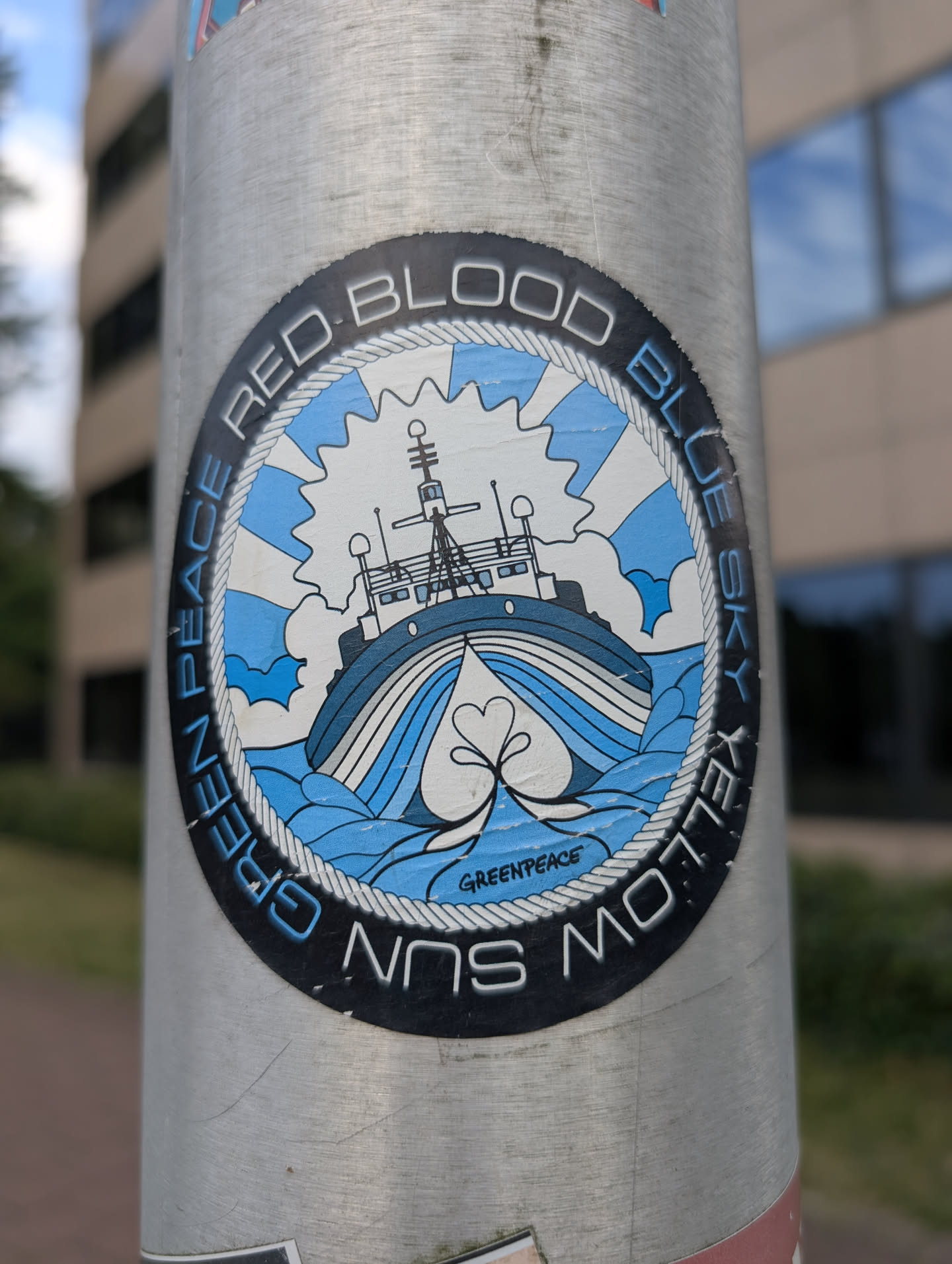 An oval sticker with a black outer ring containing the text 'RED BLOOD BLUE SKY YELLOW SUN GREEN PEACE'. Inside a rope border, a blue and white illustration depicts a ship sailing on waves under a sunburst sky, with 'GREENPEACE' written at the bottom.