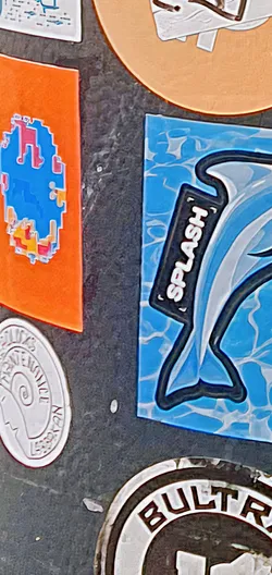 Street sticker This sticker features a dolphin swimming on a blue, wave-patterned background. The word "SPLASH" is written on a black rectangular shape above the dolphin. The sticker has a bold, outlined design, giving it a cartoonish and eye-catching appearance.