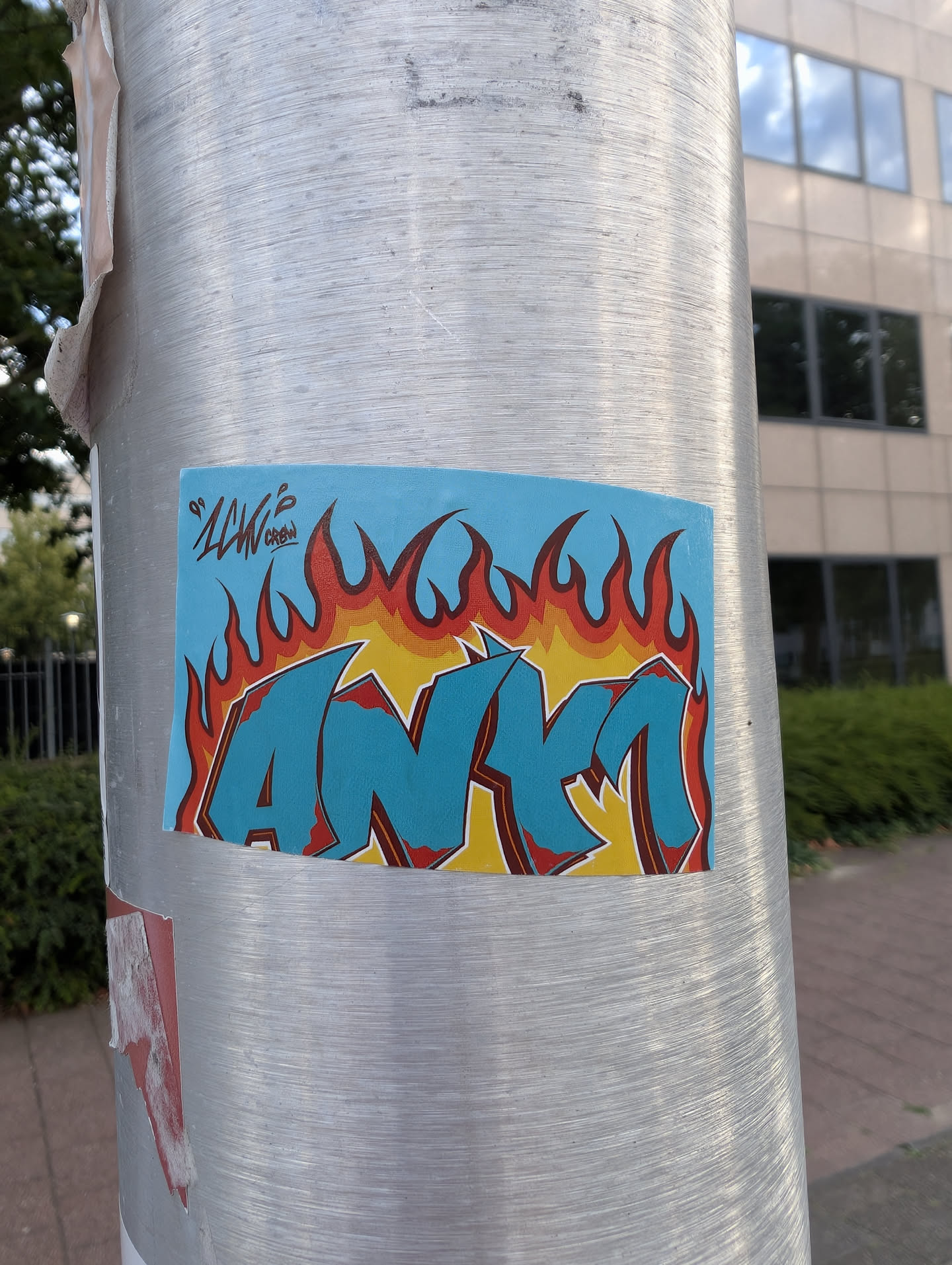 A rectangular sticker on a metal pole showing teal 'ANKM' graffiti letters with red outlines. The letters sit over yellow and red flames against a light blue background. A small 'LKW CREW' tag is in the top-left.