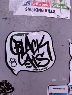 A sticker with graffiti-style text that says "BLACK CATS" and underneath in a smaller font "AND DEAD RATS". There is a small paw print symbol to the left of the main text. The sticker is white with black text and is in the shape of a speech bubble.