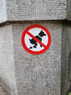 A round sticker with a red circle and a diagonal red line across a black silhouette of a dog pooping.  The sticker is affixed to a grey stone pillar.