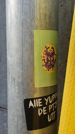 Street sticker A green sticker with an abstract design in shades of yellow and purple. Below it, another sticker with white text on a black background. The text is in a stylized font and appears to be in a language other than English. The style is reminiscent of punk or alternative culture.
