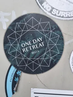 Round sticker with a dark background featuring a geometric pattern and fern leaves.  The text "ONE DAY RETREAT" is prominently displayed, with "IBIZA" written underneath. A portion of a light blue sticker is visible underneath, with the word "FACEBOOK" partially visible.