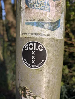 Street sticker The sticker is round with a black background and white text and symbols. It reads "SOLO" at the top, followed by three "X" symbols stacked vertically, and "AMSTERDAM" at the bottom. The sticker is affixed to a weathered metal pole.