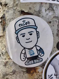 Street sticker Round sticker featuring a cartoon character wearing a hat with 'CHLOF' written on it. The character has a simple design and is wearing a t-shirt with a small logo. The sticker has a slightly worn look, suggesting it has been placed outdoors for a while. The background is a weathered surface, possibly a wall or pole.