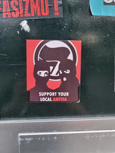 Street sticker A sticker featuring two masked figures and the text 'Support Your Local Antifa'. The sticker uses a bold red and black color scheme and has a simple, graphic design. 