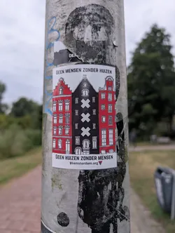 Street sticker The sticker features a stylized depiction of three Dutch canal houses. The leftmost and rightmost houses are red, with white detailing for windows and doors. The central house is black with three large white 'X' symbols on its facade, and smaller white windows. The houses have characteristic stepped gables. Above the houses, text reads 'GEEN MENSEN ZONDER HUIZEN' (No people without homes). Below the houses, the text 'GEEN HUIZEN ZONDER MENSEN' is present, followed by a website address and a small red triangular logo.
