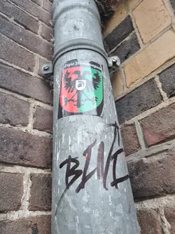 A sticker affixed to a metal pipe, showing a coat of arms with a double-headed eagle. The text above the crest reads 'Legio Noviomagi'. Below this sticker, there's some graffiti that says 'BLVC'. The pipe is set against a brick wall.