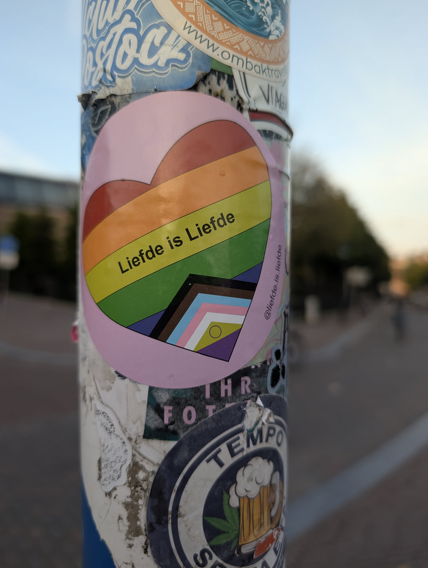 A circular pink sticker displaying a heart filled with the Progress Pride flag. The text 'Liefde is Liefde' is written across the center, and the Instagram handle '@liefde.is.liefde' is printed on the right side.
