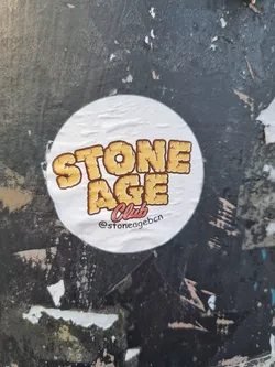 Street sticker Round sticker with the text "STONE AGE Club" in a vintage style font. The text is yellow/gold with a rocky texture. The background of the sticker is white and there is a social media handle on the bottom. The sticker is affixed to a dark gray, textured surface.