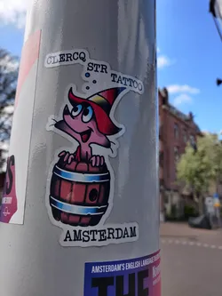 Street sticker A sticker featuring a cute cartoon character with a wizard hat sitting inside a wooden barrel. The text 'GLERCQ STR TATTOO' and 'AMSTERDAM' are visible on the sticker.